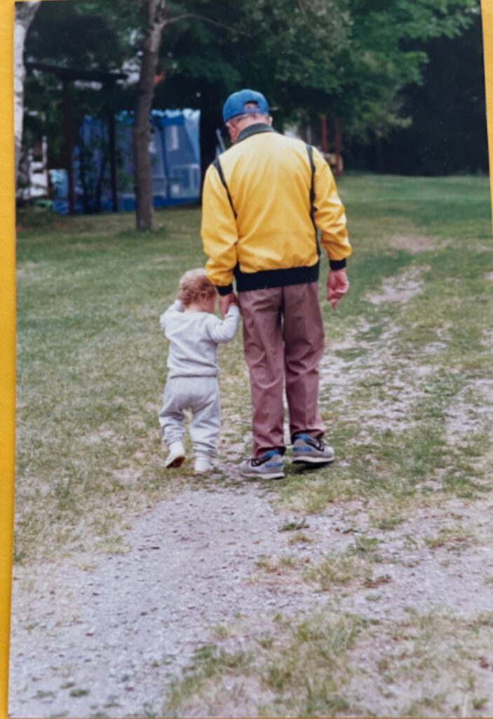 A father and child walk hand in hand along a Camp Naivelt path