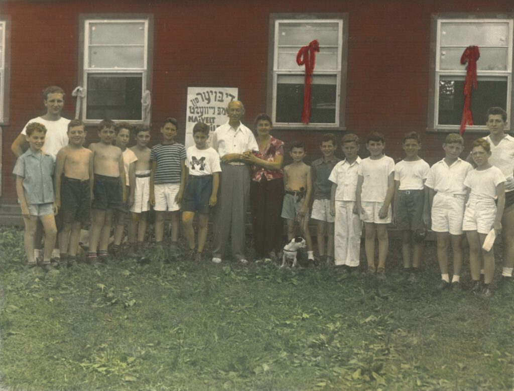 Children gather with a Yiddish harvest (Sukkes) sign outside a camp cabin